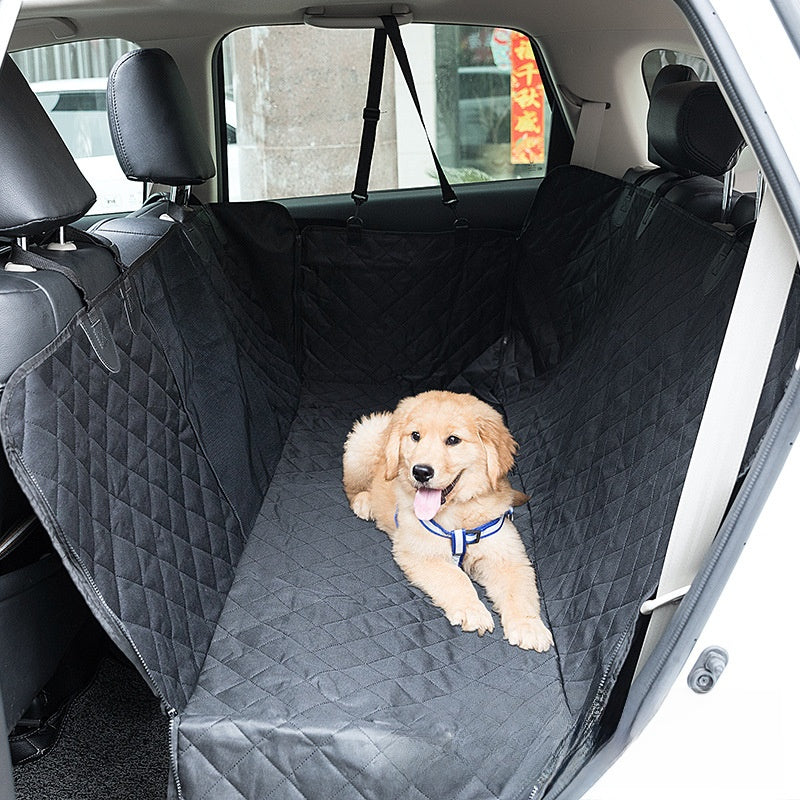 Dog sitting on a black quilted car seat cover in the back of a vehicle.